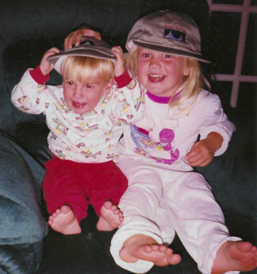 Young Blakely and her even younger sibling wearing Cisco hats sitting in a chair together.