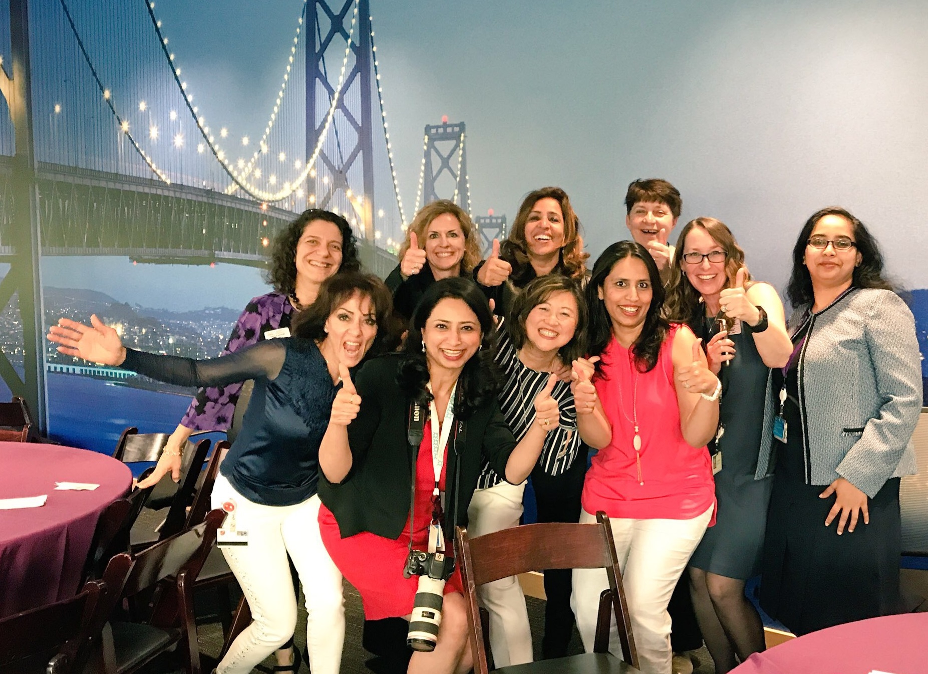 Varsha, with a camera around her neck, and her peers give a thumbs up in front of a mural of a bridge.