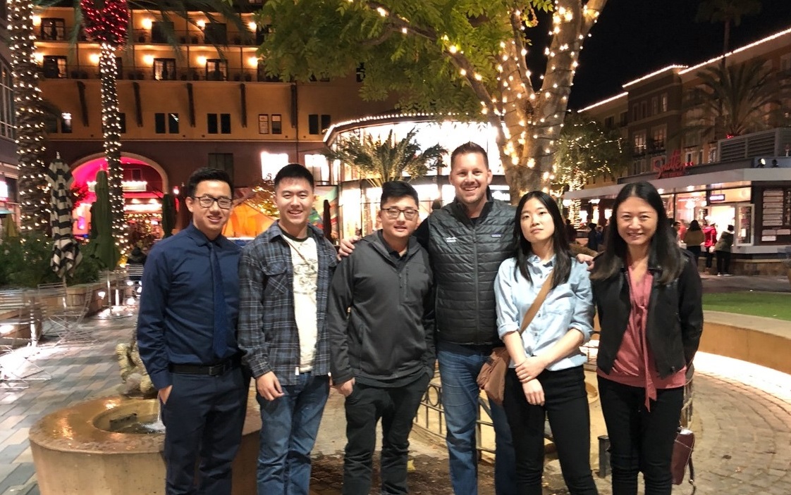 Peter and his peers smile with their boss Bryan surrounded by palm trees decorated with lights.