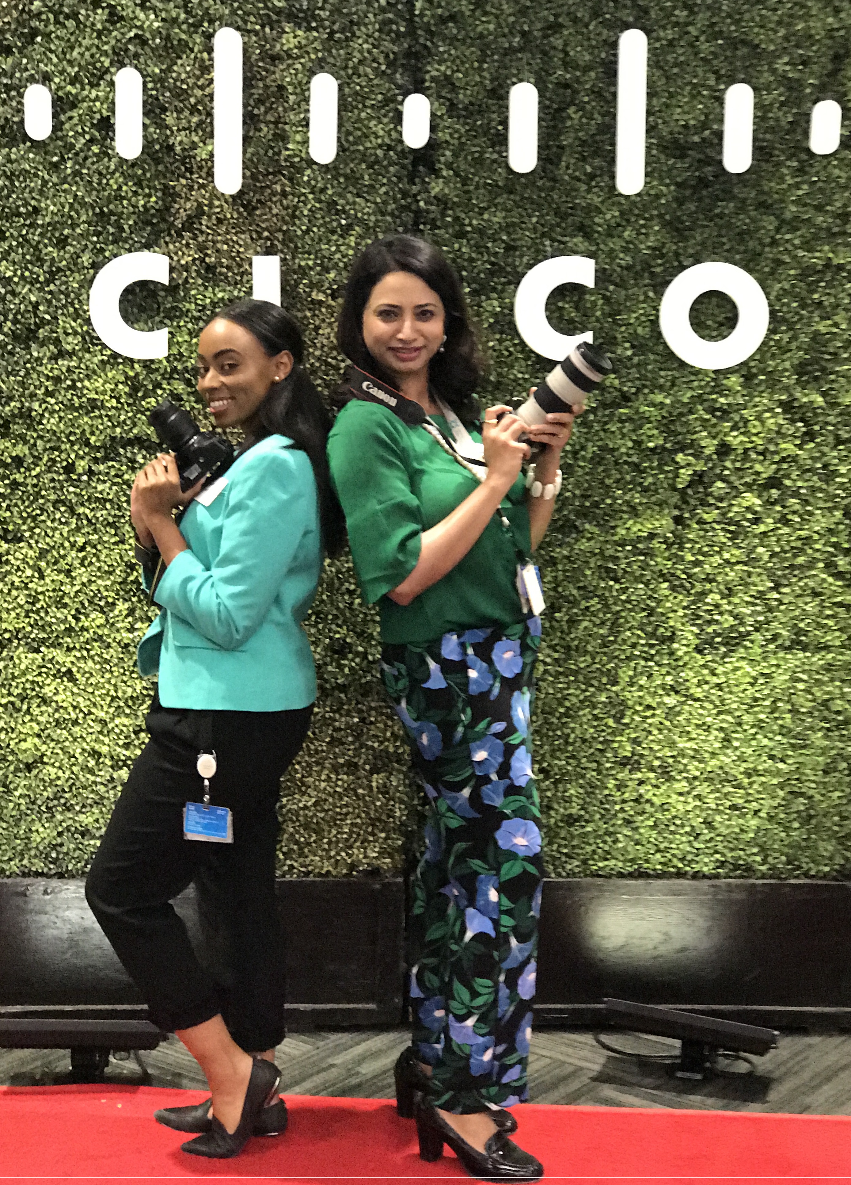 Varsha and a peer stand back to back smiling with their cameras in front of a topiary wall with a Cisco logo.