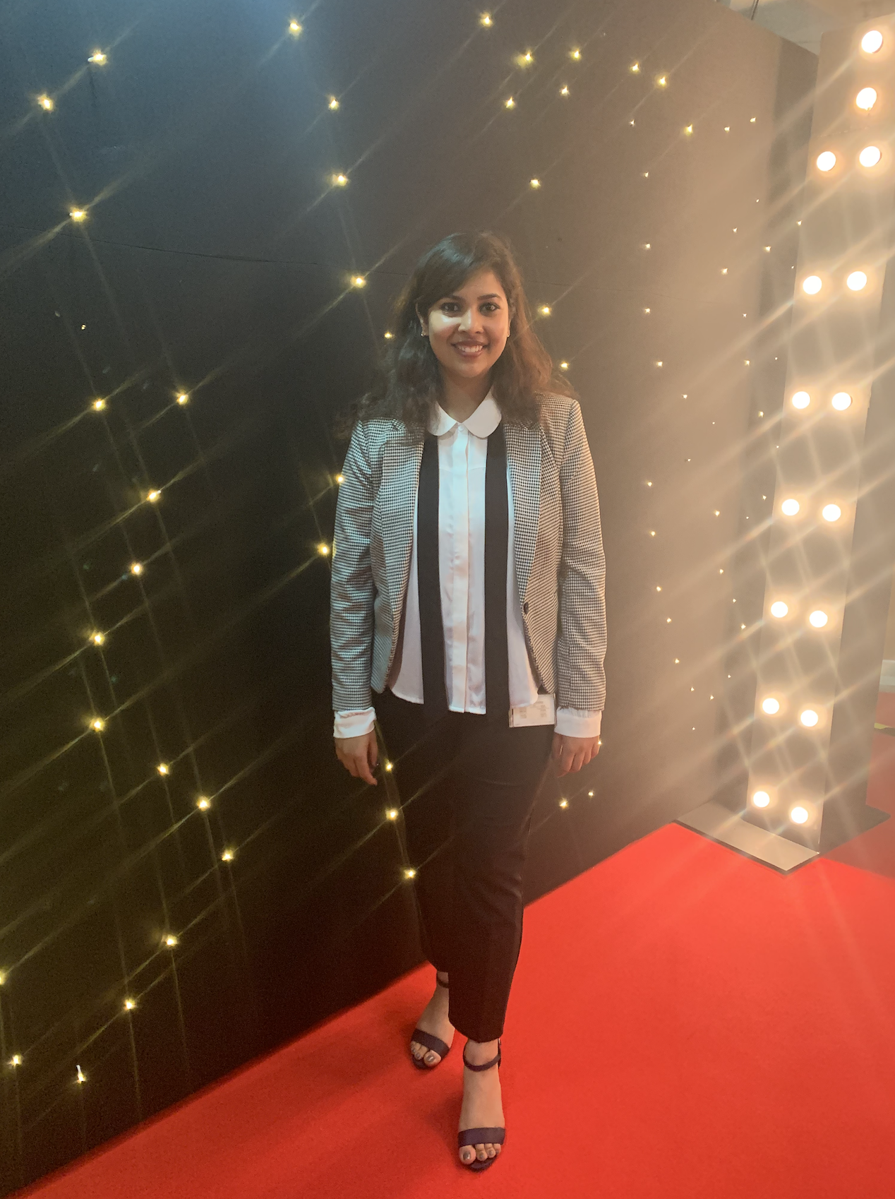 Nikita stands in front of a wall of lights at an event on top of a red carpet.