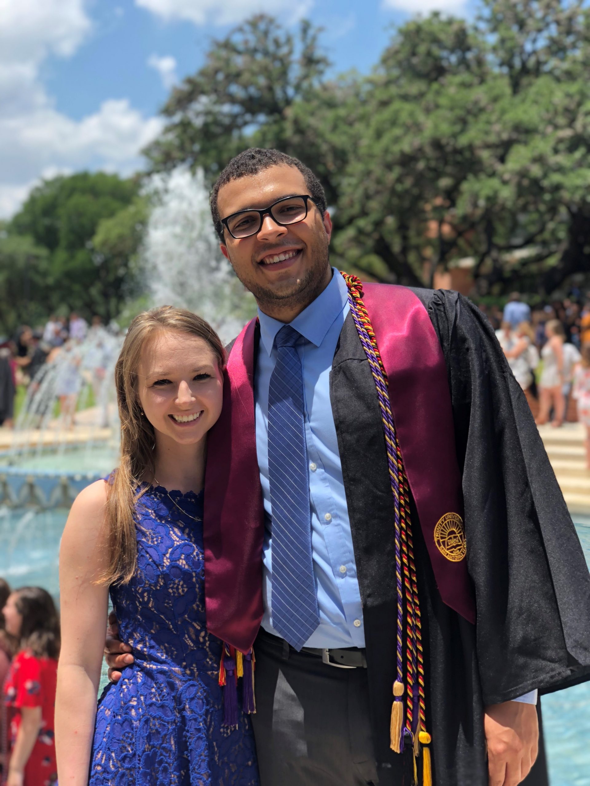 Julian stands proudly in front of a fountain wearing his graduation gown.