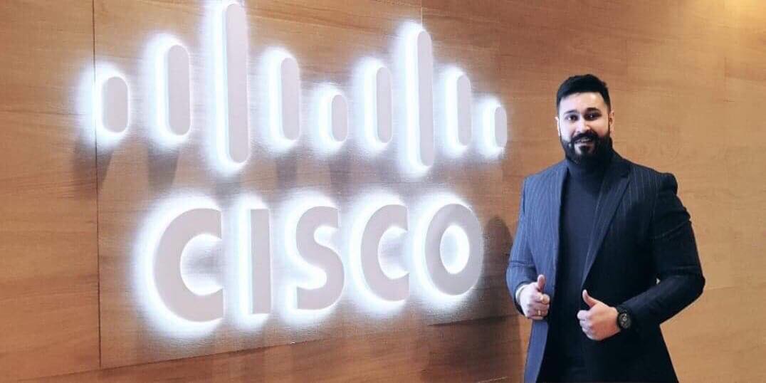 Joy smiles gripping at his blazer next to a light up Cisco sign.