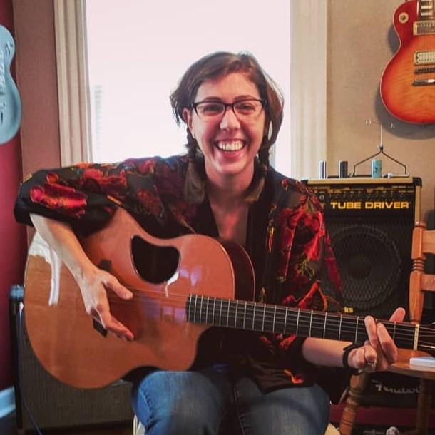 Annie has a huge smile as she holds a guitar surrounded by other guitars and speakers.