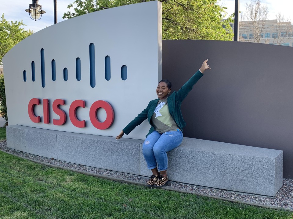Amber smiles with her arms pointed out beside her sitting on a Cisco sign outside of a Cisco campus.