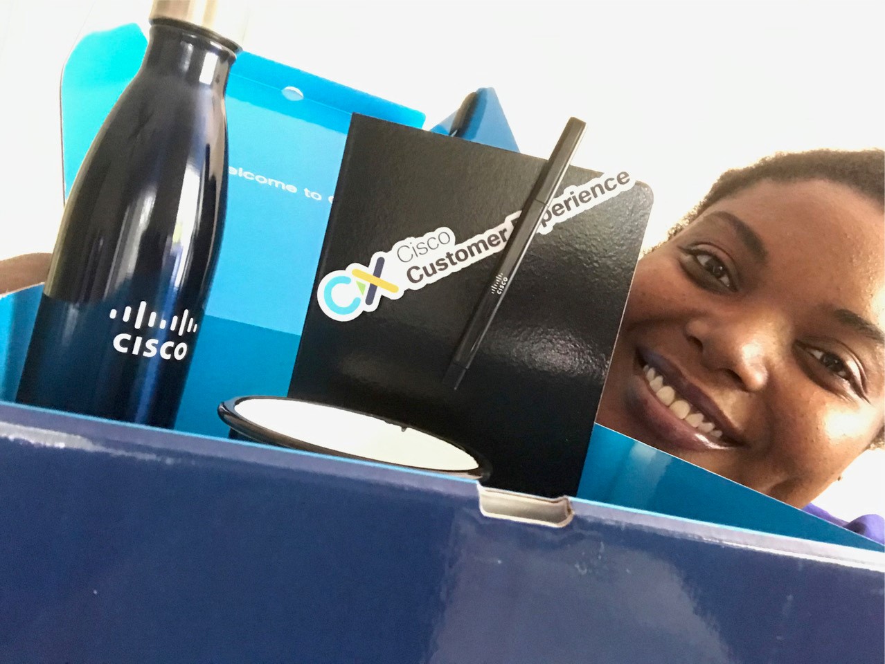 A Cisco Customer Experience new hire smiling with their new-hire swag box, complete with water bottle, mug, and a notebook.