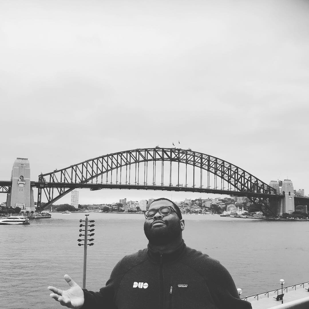A black and white photo of Karl wearing a DUO branded jacket stands overlooking a bridge in Australia.