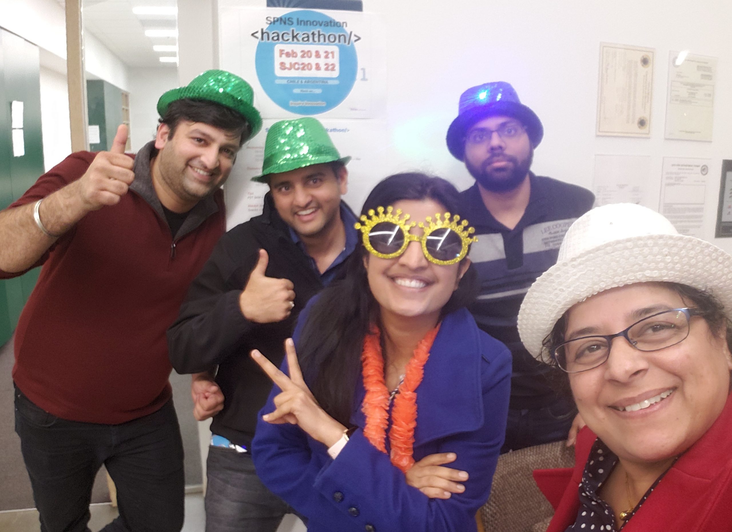 Rashmi and 4 of her peers wear silly hats and glasses under a SPNS Innocation Hackathon poster.
