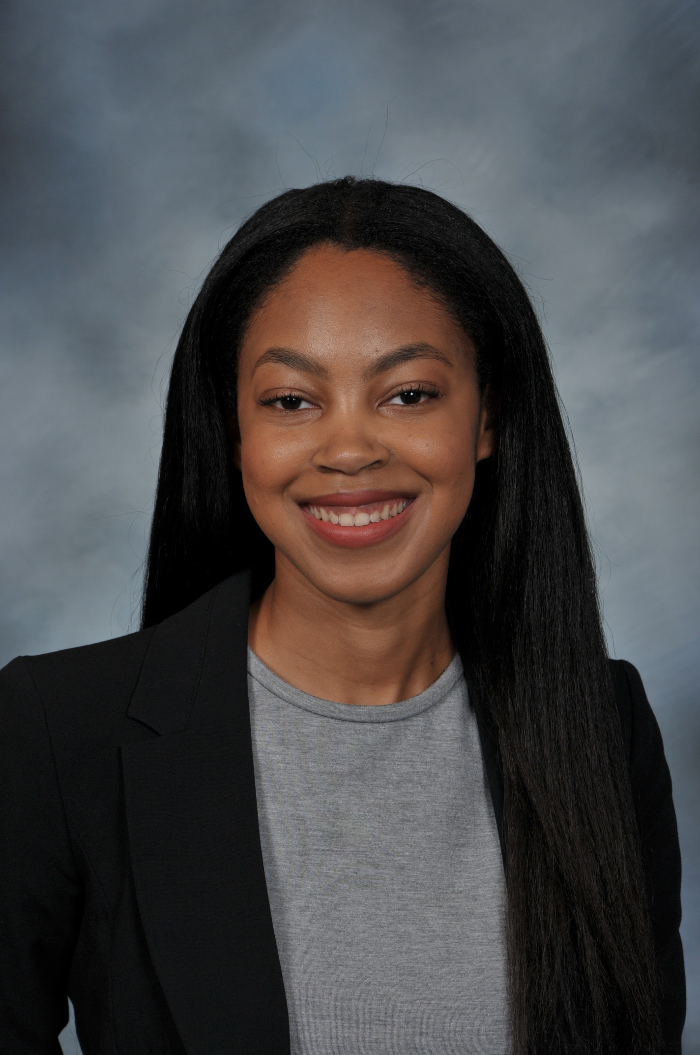Azhia smiles in front of a gray backdrop wearing a black blazer.
