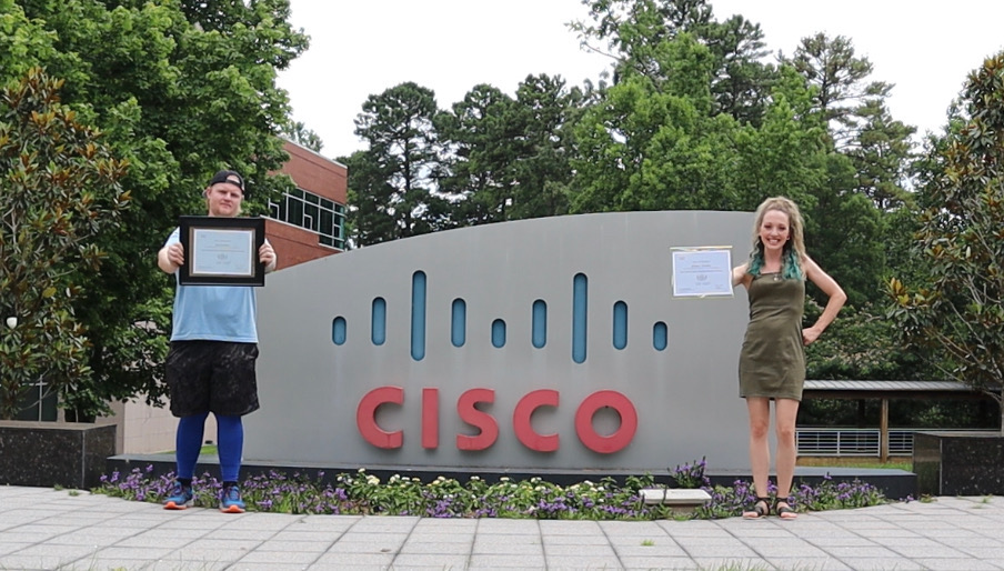 Miranda and her husband holding up their certificates in front of a Cisco sign outside.