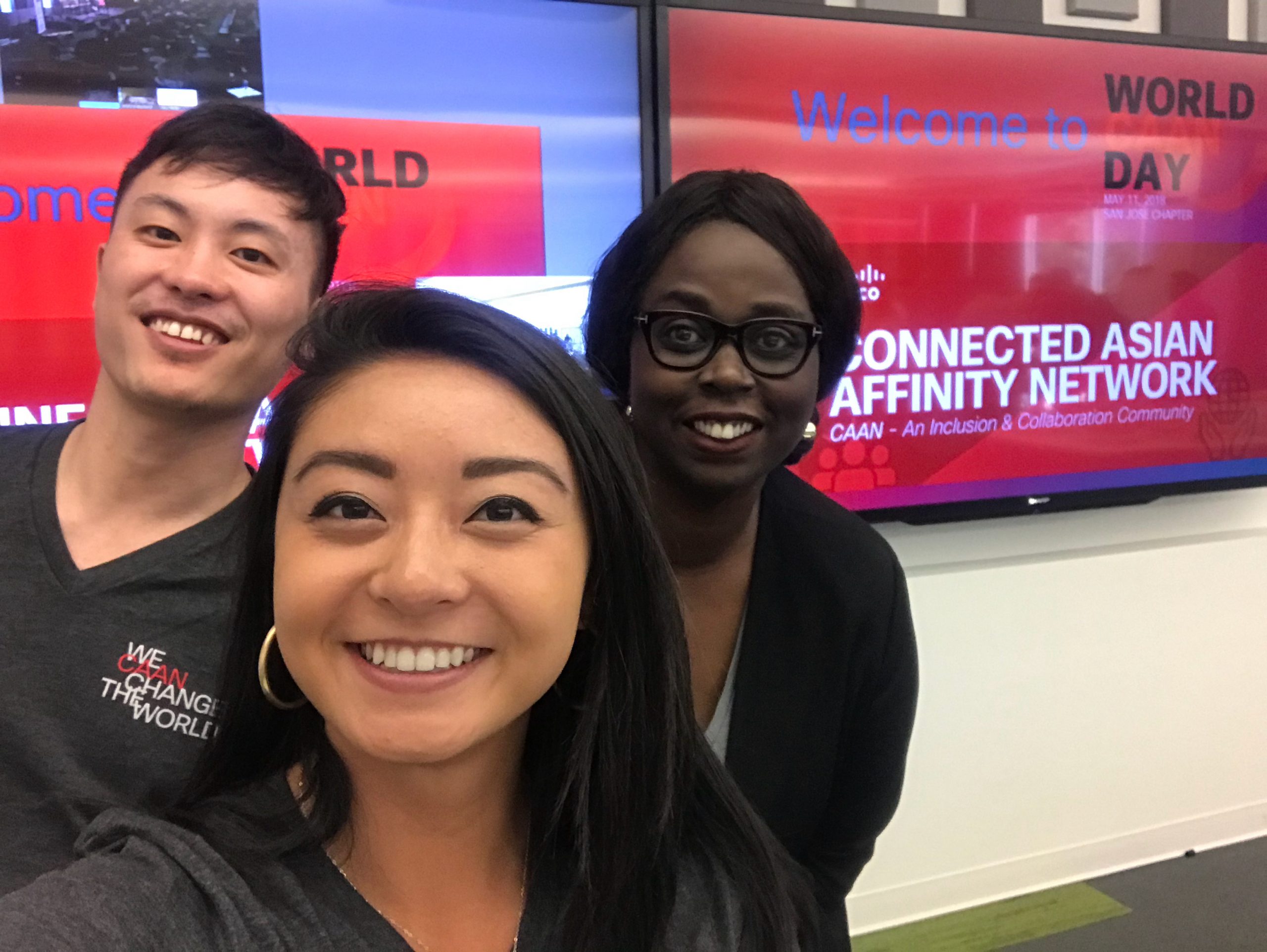 Melissa smiles with two of her peers in front of two tv's with the Connected Asian Affinity Network logo displayed.