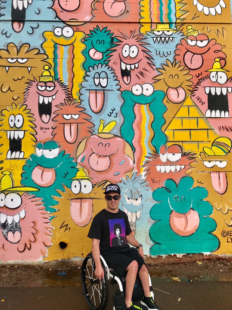 Hideaki wears a hat and sunglasses in front of a colorful mural.