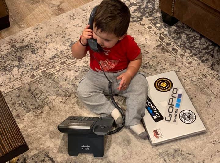 Nic's youngest son sits on the ground next to a laptop holding a Cisco phone to his ear.