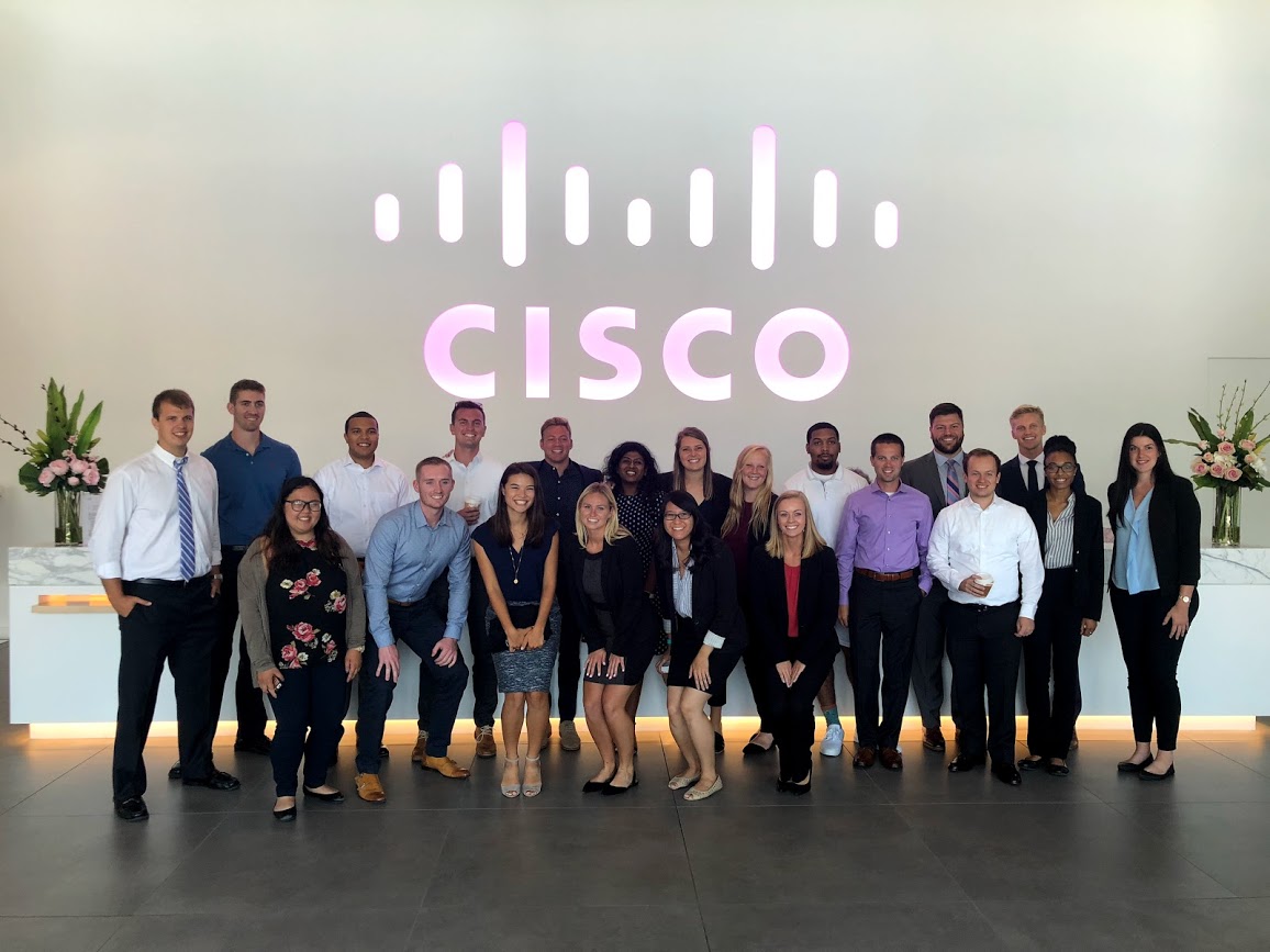 Prabha's Cisco LIFT Class in a Cisco lobby with a pink light up Cisco sign behind them.