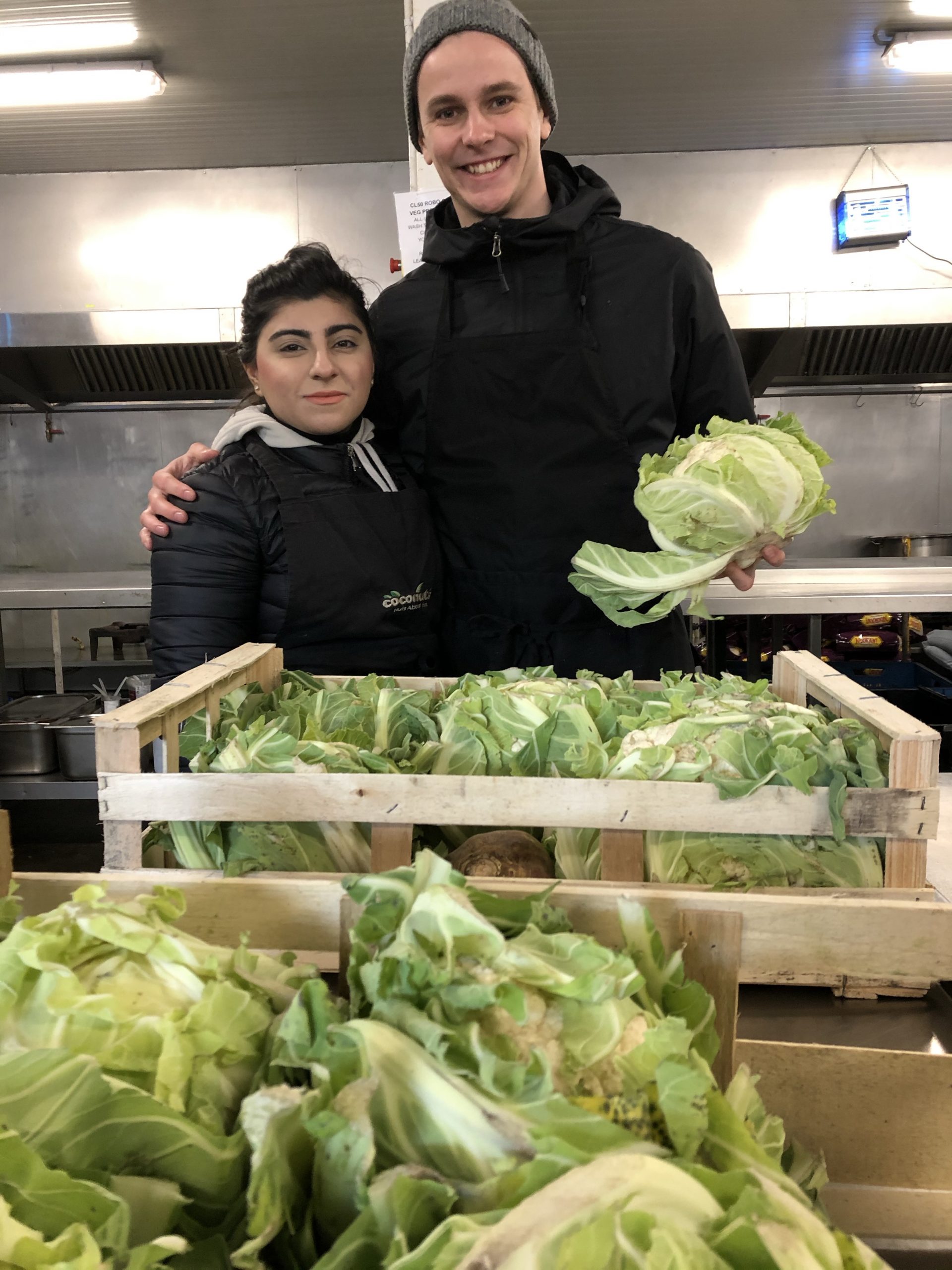 Nico stands with his peer behind boxes of lettuce in The Jungle's kitchen area holding a head of lettuce.