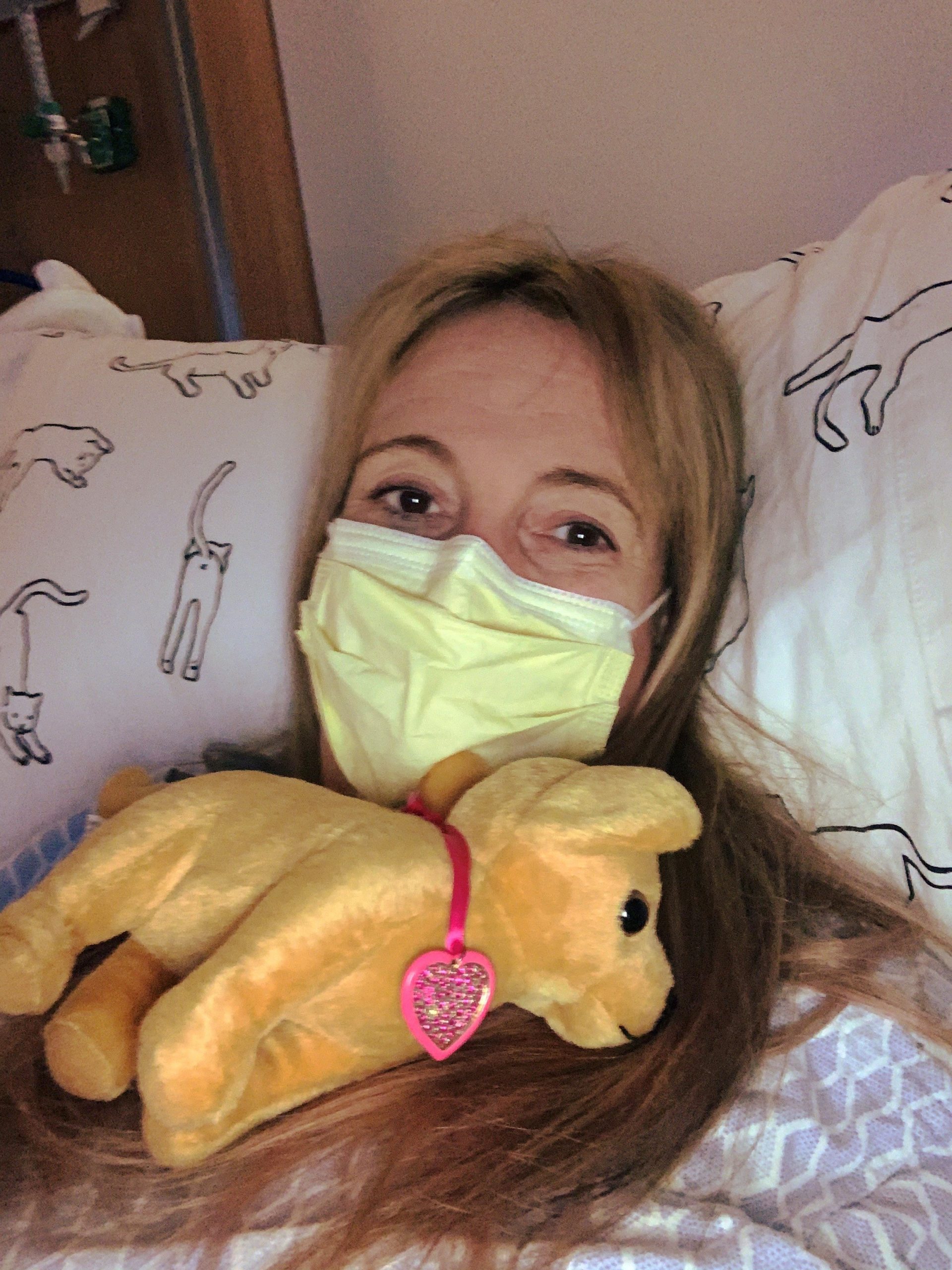 Rachael wearing mask with a stuffed animal while laying in hospital bed