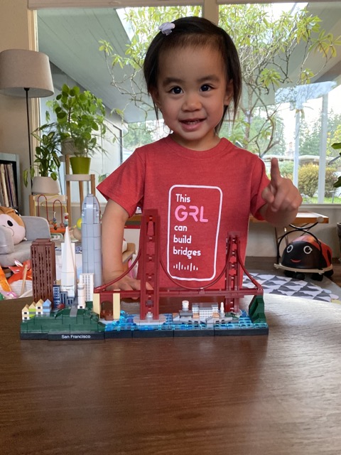 Young asian girl wearing red t-shirt that says "This girl can build bridges" with a Cisco logo, building Golden Gate bridge with legos holding up her fingers with a number 1.