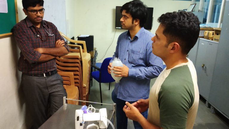 Three men talking in front of a ventilator
