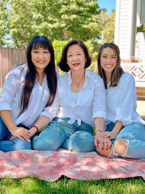 Renee, her daughter, and mother sit on a blanket, holding hands and smiling in white shirts and jeans.