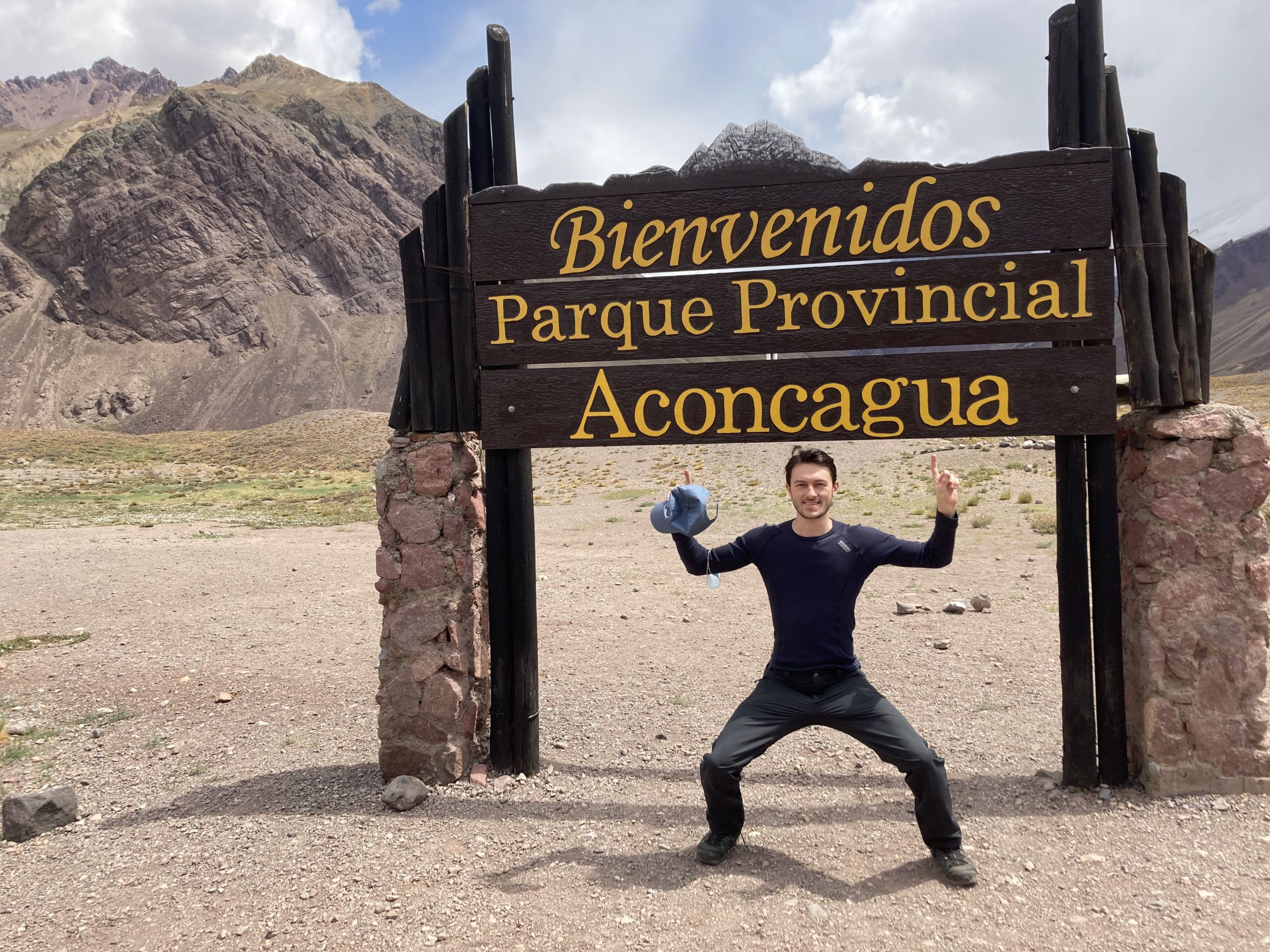 Jeremy stands under sign pointing up at it that reads bienvenidos parque provincial Aconcagua.