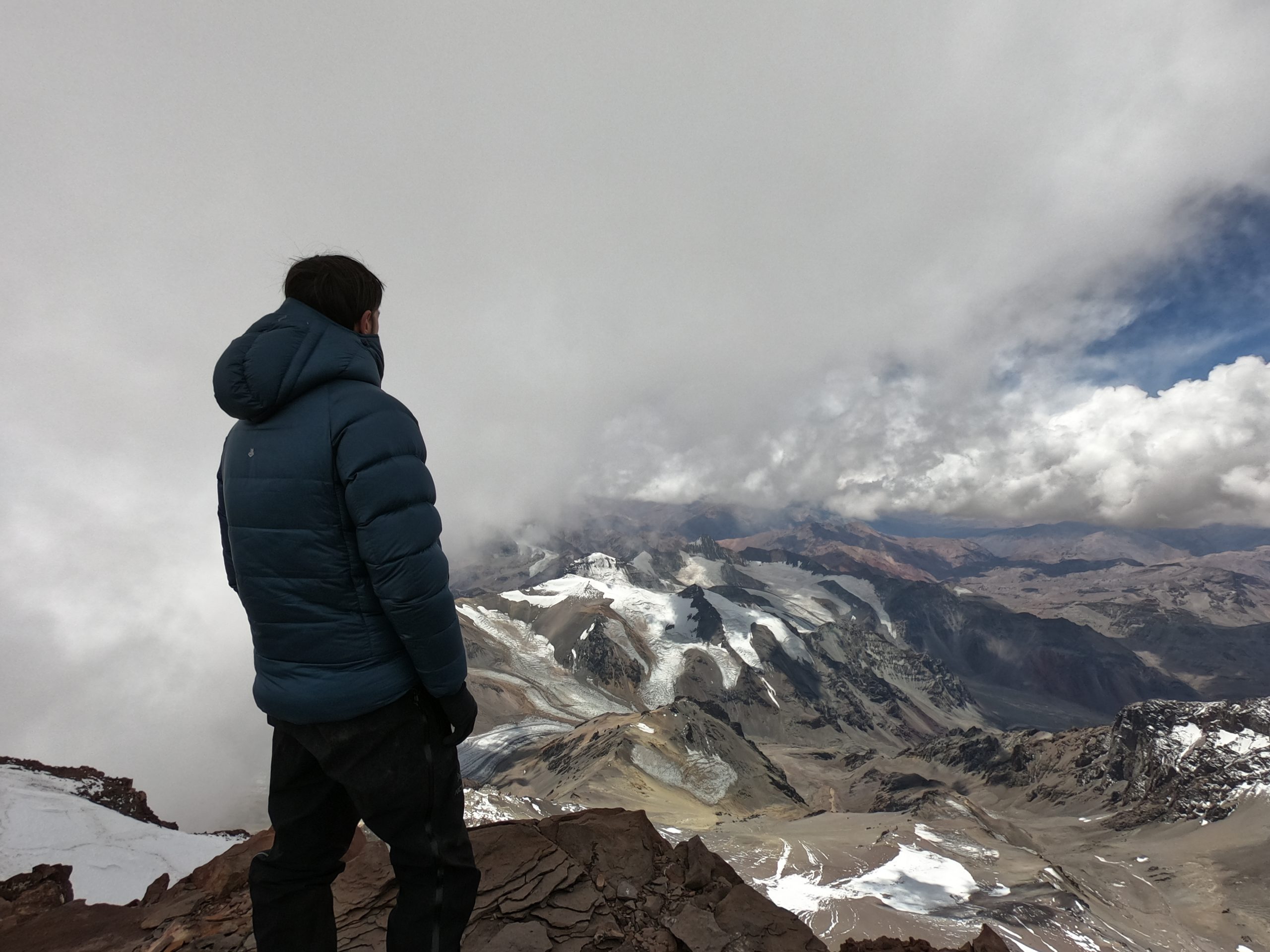 Jeremy stands with back facing camera in blue puffer jacket looking down into mountain valley.