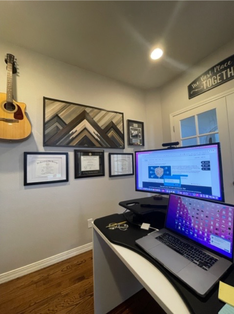 Work from home office with guitar and diplomas on the wall and desk with external monitor and laptop.