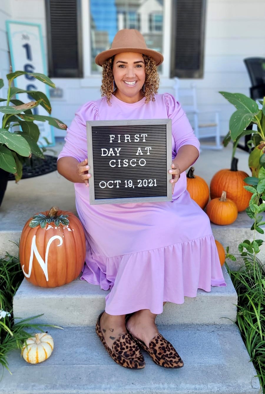 Jessica Norwood sits on porch steps holding a sign that reads, "First Day at Cisco Oct 19, 2021." She is wearing a pastel pink dress and is surrounded by pumpkins.