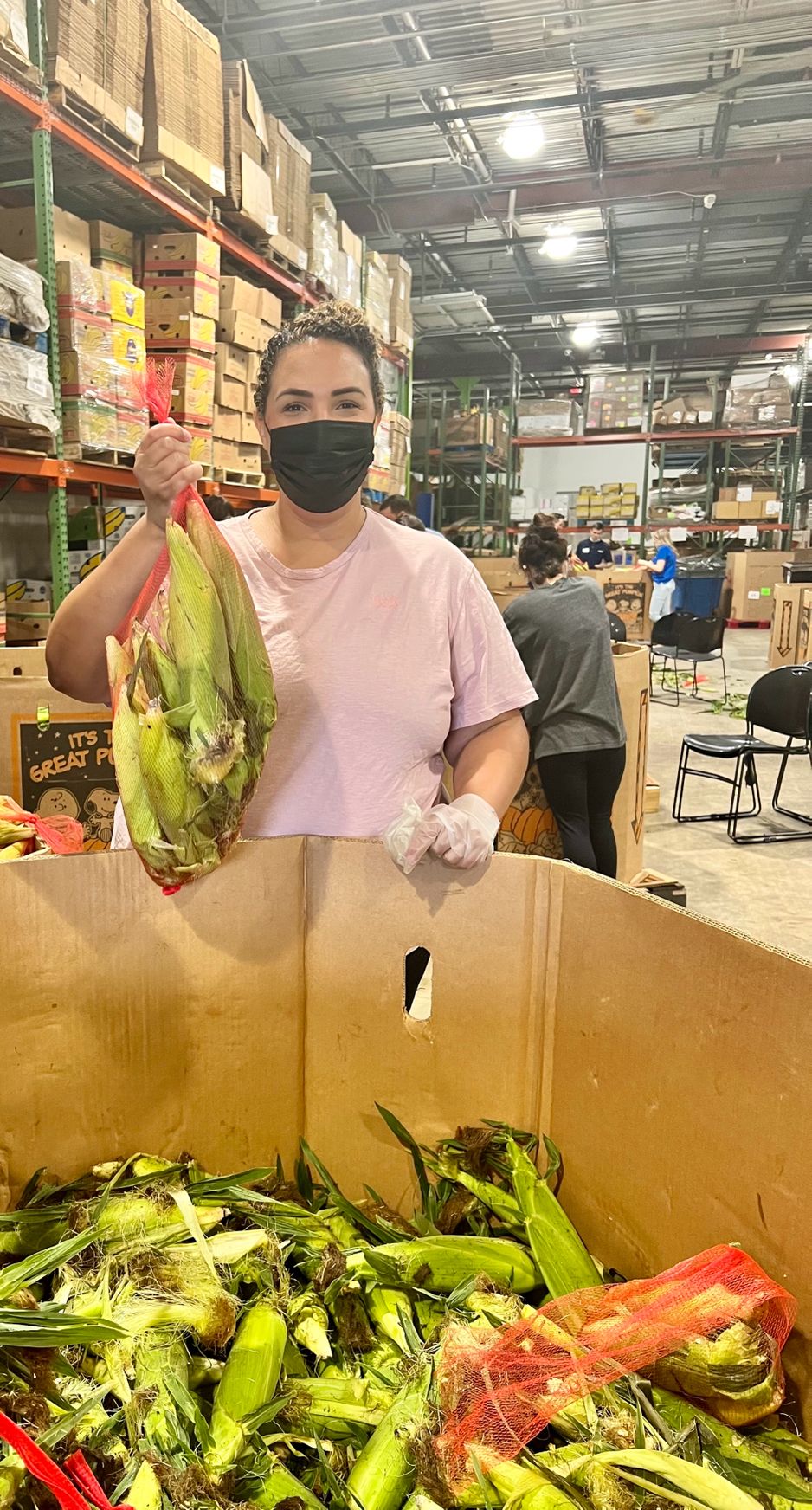 Jessica Norwood wears a face mask and holds up a bag of unhusked corn. She is in a warehouse and there is a large box of unhusked corn in front of her.