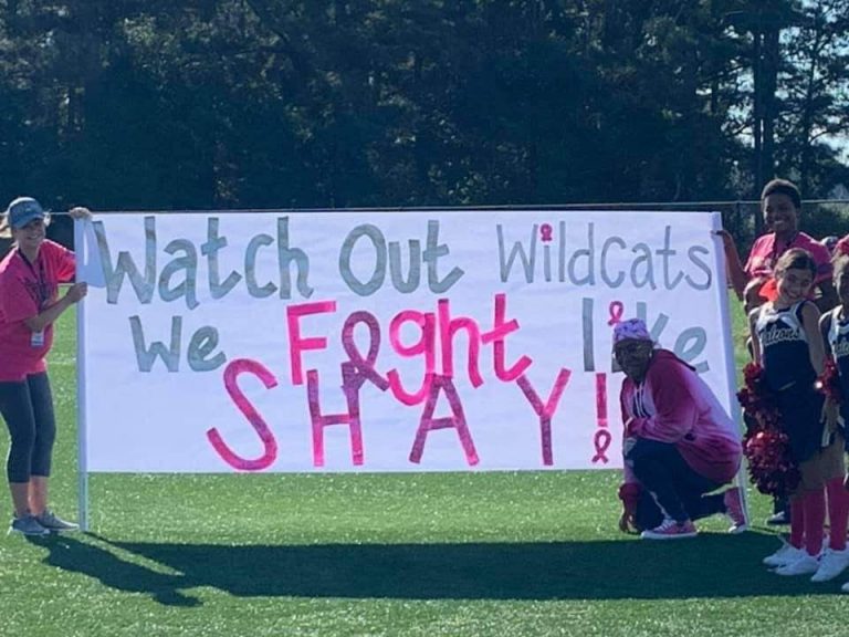 People huddle around a large sign that reads "Watch out Wildcats we fight like Shay!"