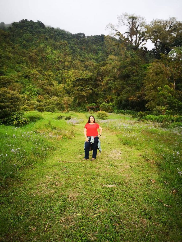 A woman in a red top standing outside with lush green grass and trees around her