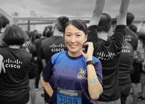 Crowd in the back in black and white with backs to the camera wearing Cisco logo shirts and girl in the foreground in color holding up a "C" with her hand.