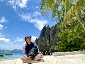 Woman sitting cross-legged on the beach holding Cisco backpack looking up at palm tree.