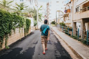 Man with teal backpack and red shoes walking with back towards the camera down an alley.
