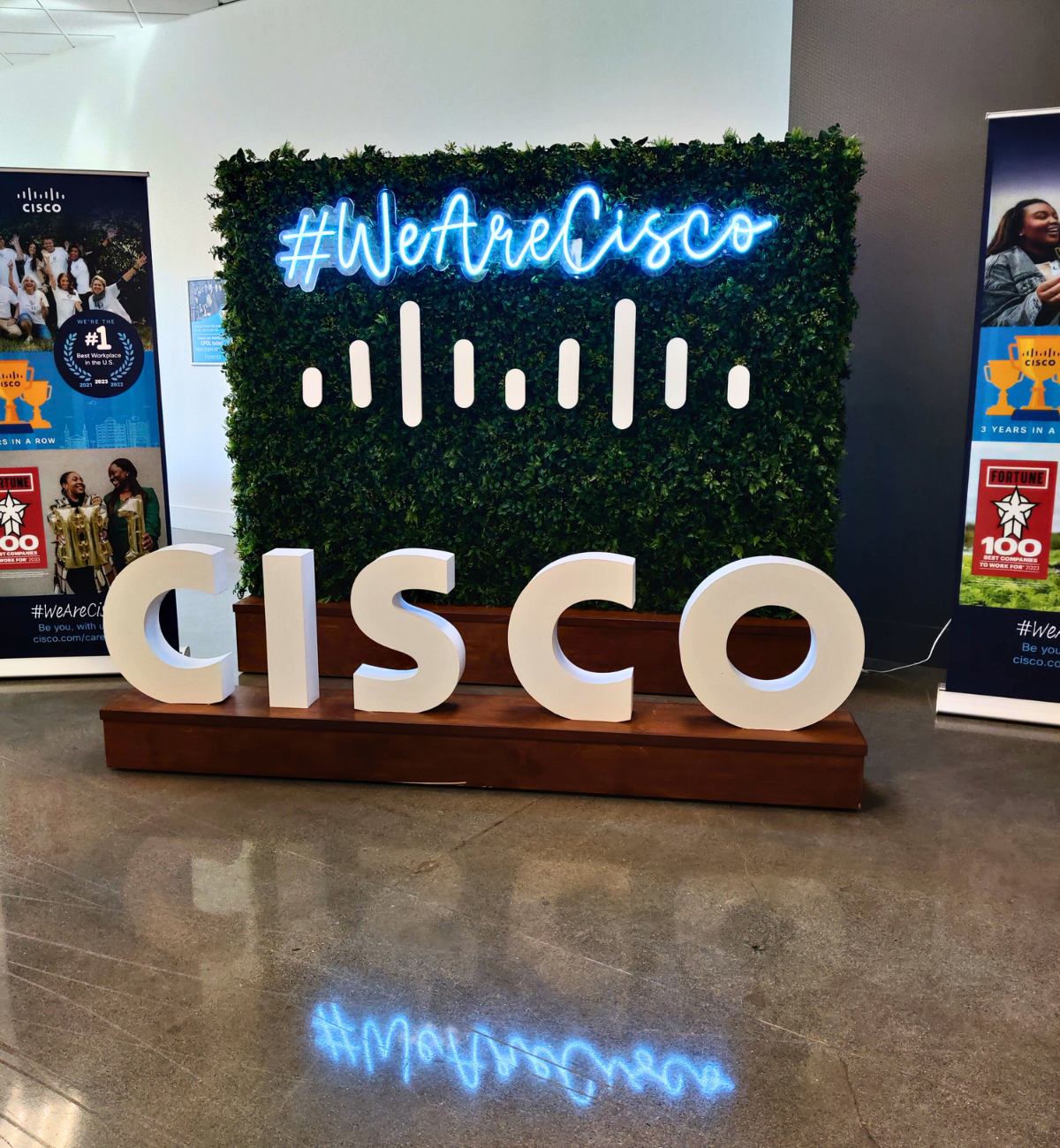 Large sign that reads "#WeAreCisco " and "Cisco" with faux leaves in the background.