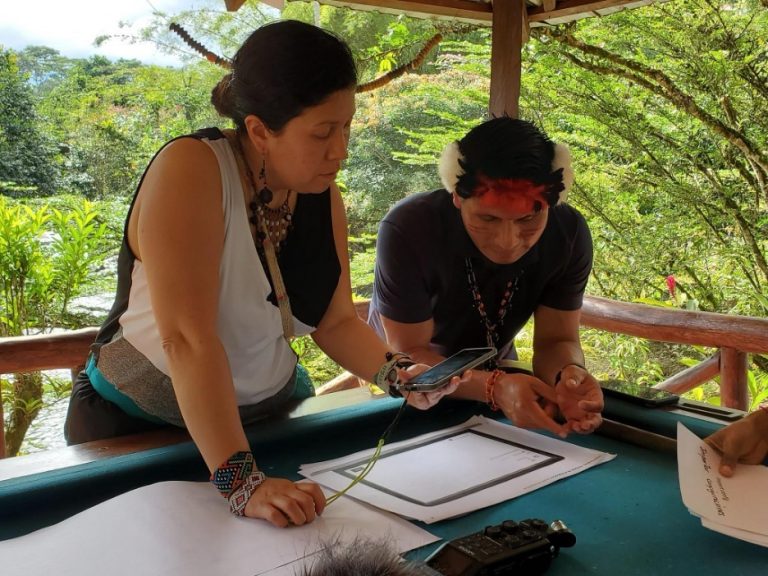 Two people looking at document together, with one holding a phone above the document with green trees behind them