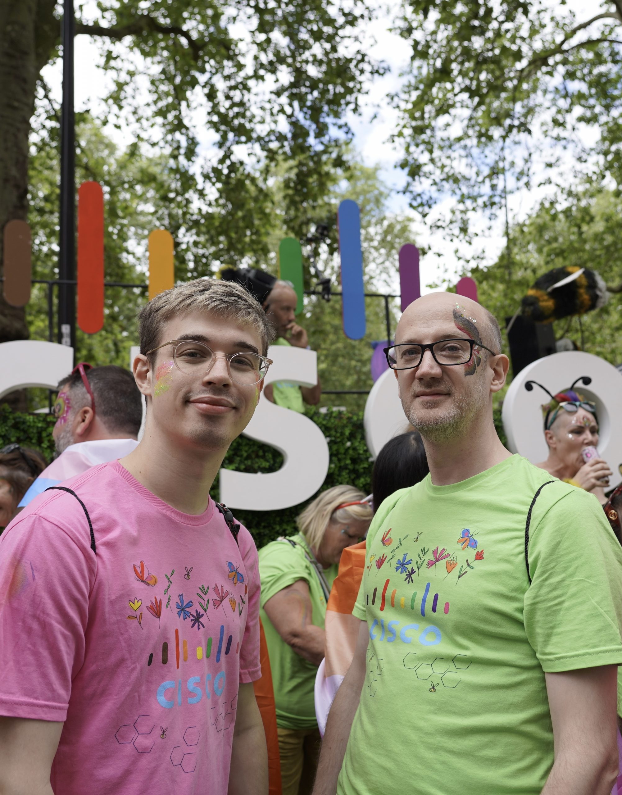 Two caucasian males wearing glasses, one in a pink t-shirt, one in a green t-shirt, standing in front of a big rainbow-logo Cisco pride parade float.