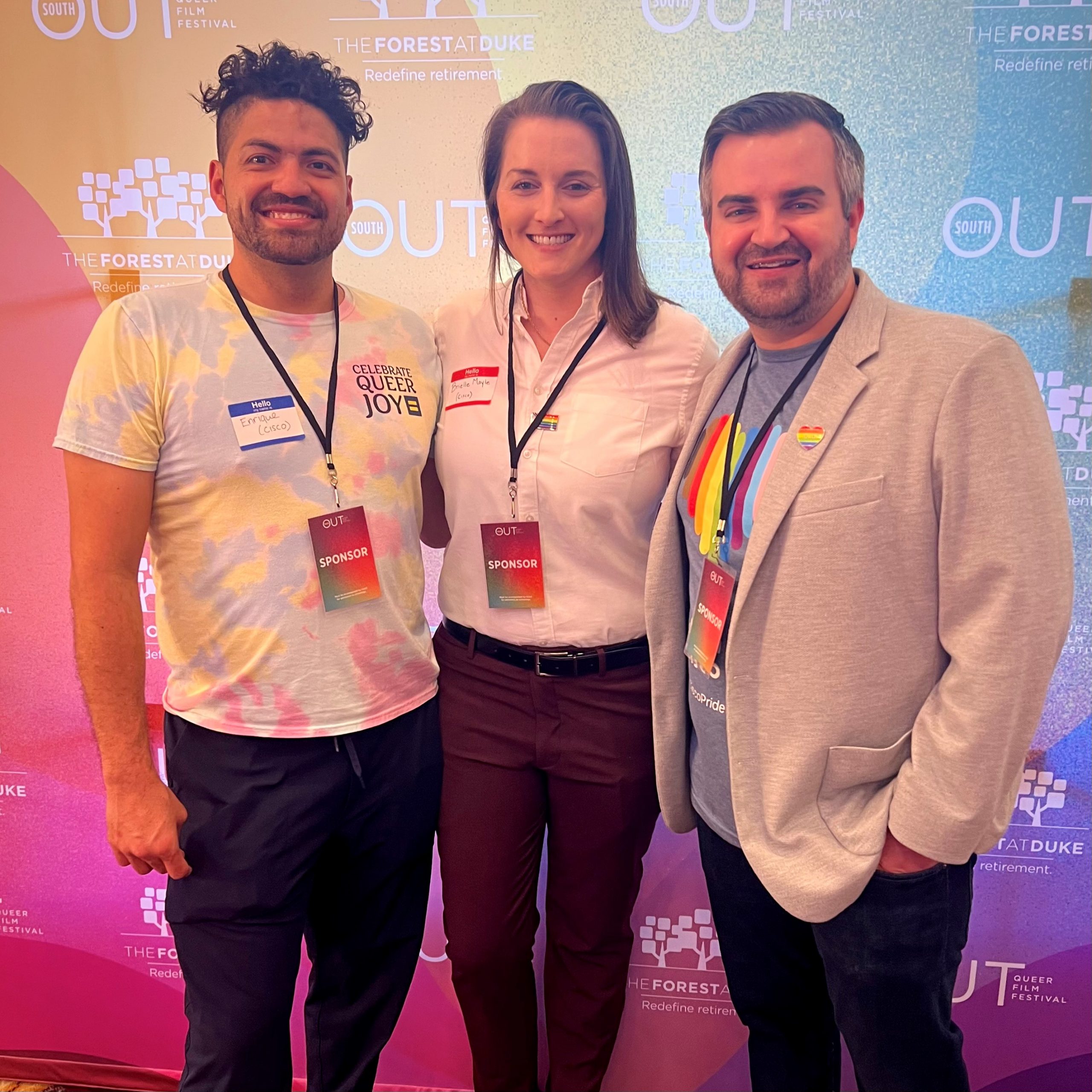 Two men and a woman smiling at camera with arms around each other standing in front of step and repeat wearing lanyards with name tags.