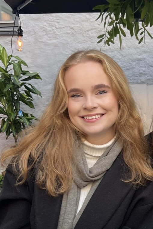 Caucasian woman with blonde hair smiling at the camera. She is wearing a cream turtleneck, gray scarf, and black coat.