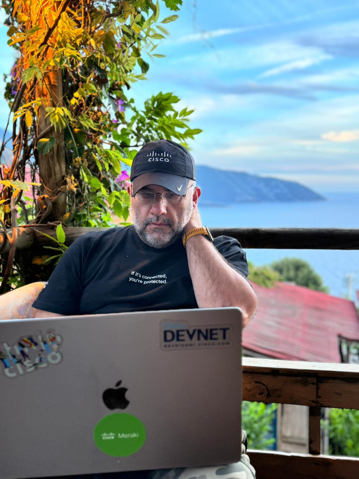 Caucasian male wearing black Cisco baseball cap working from laptop in tropical-looking location. 