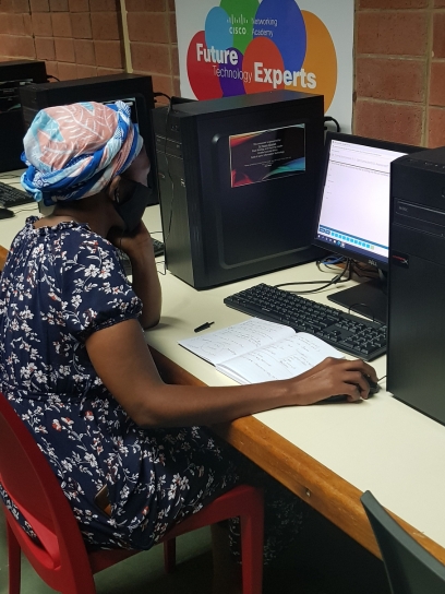 A young African woman wearing a black face mask, dark blue floral dress, and a blue headscarf sits at a desk working on a desktop computer.
