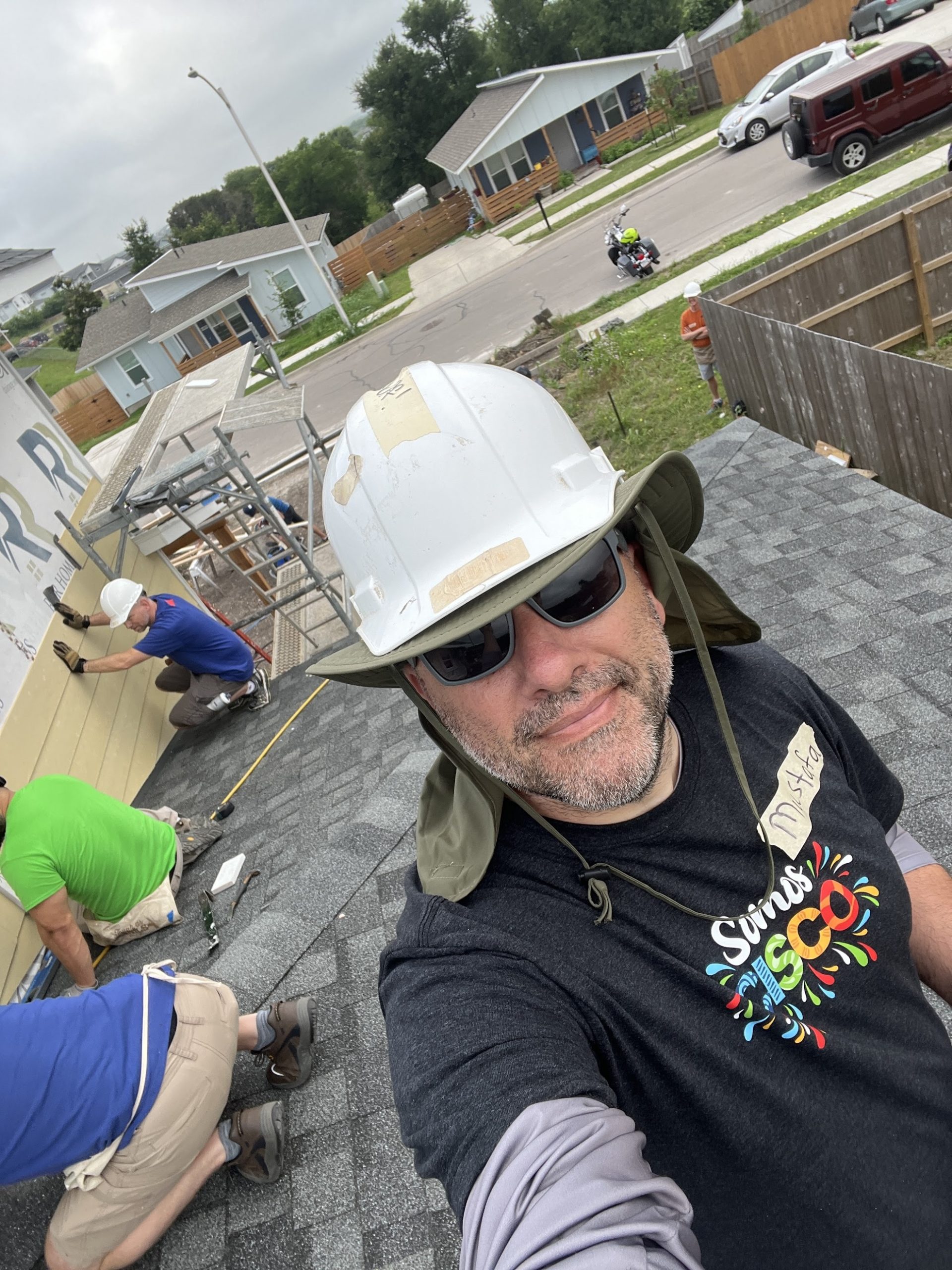 Caucasian male wearing hardhat, sunglasses, and "somos Cisco" t-shirt at a construction site taking a selfie.