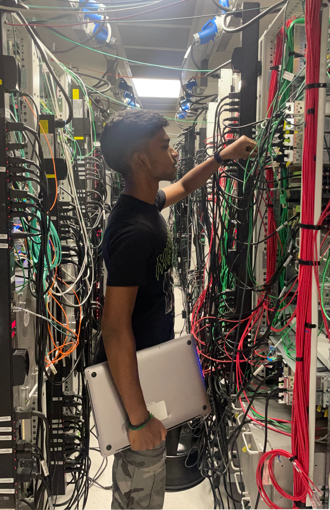 A medium-skinned man with dark brown hair stands in an aisle full of hanging cables holding a laptop. 