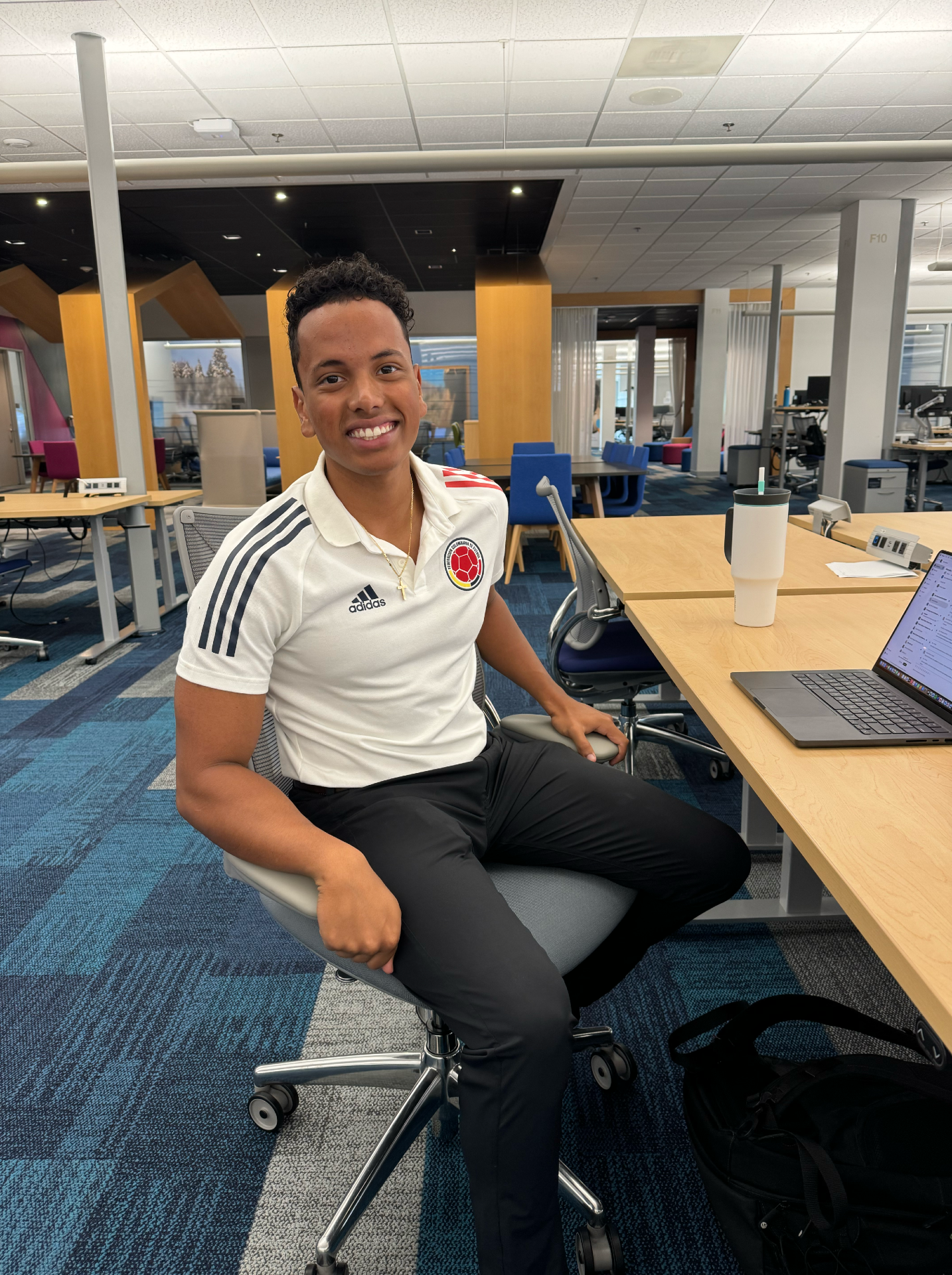 Tan-skinned young man with brown hair and brown eyes wearing white athletic shirt with red stripes at the shoulders and black pants sits at desk with laptop open, smiling at camera with open office in the background. 