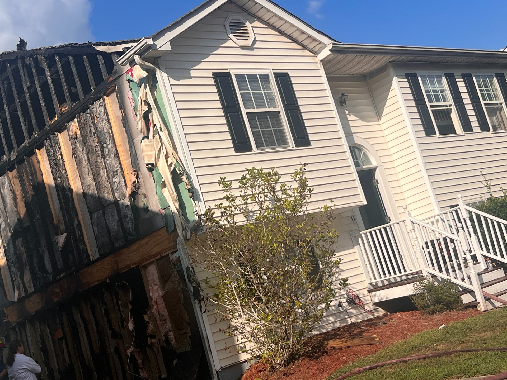 Cream-colored two-story house with black shutters. The left side siding is removed and wood is exposed from fire damage.