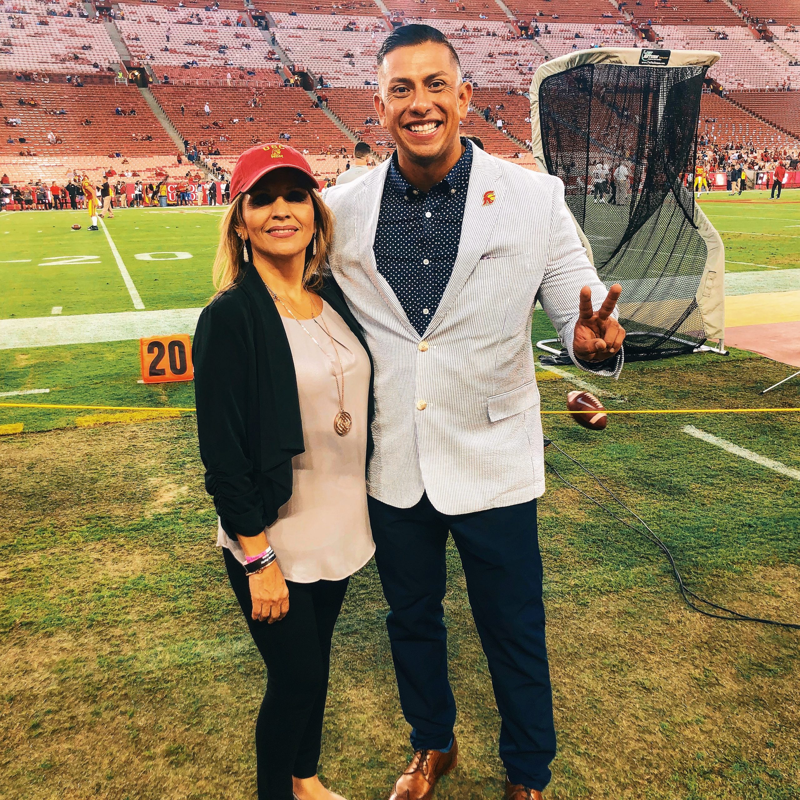 A woman in black pants and blazer, cream top and red baseball cap and man in seersucker blazer and polka dot shirt and black pants holding a peace sign stand on the sidelines of a football field.