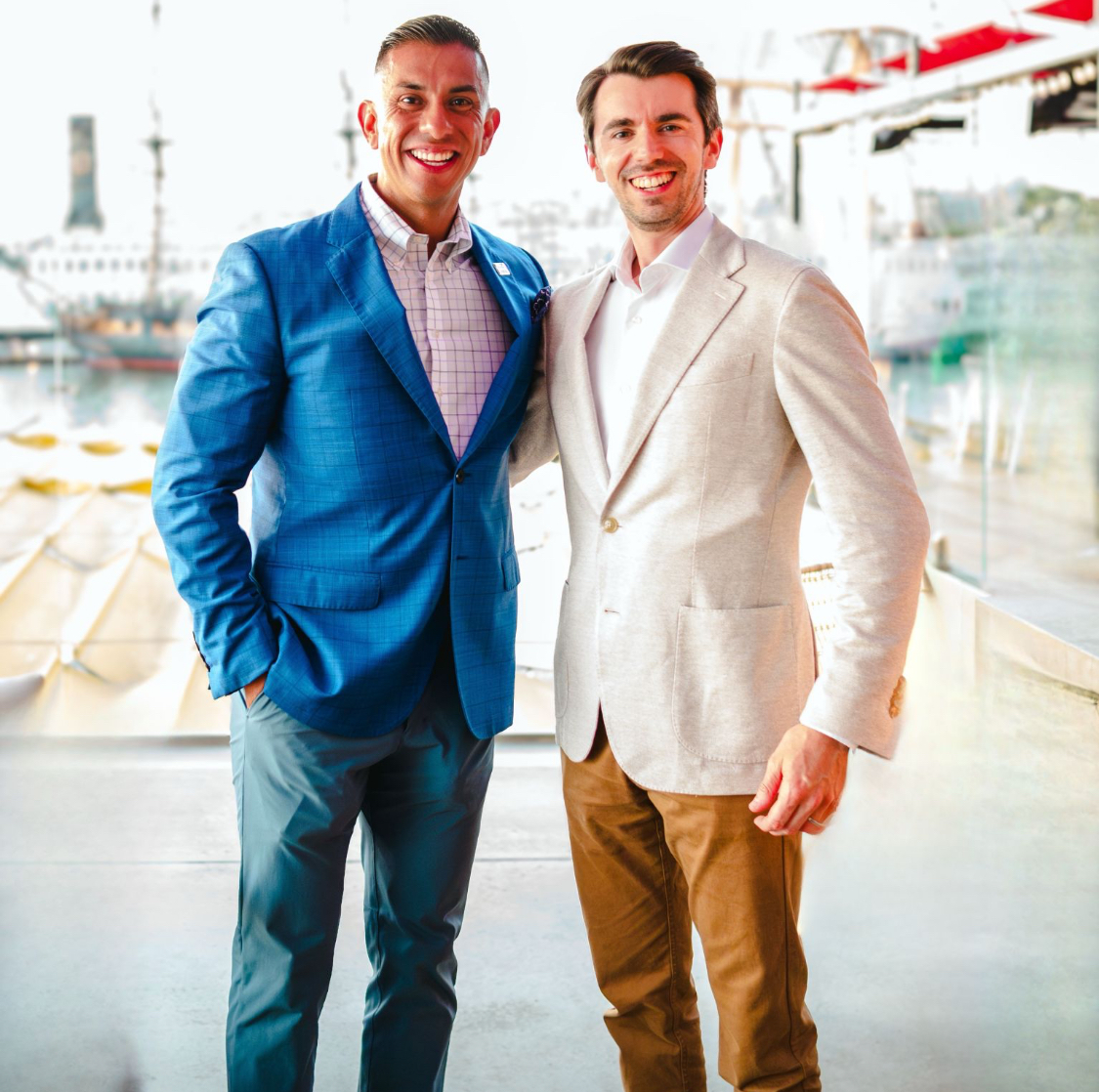 Man in blue blazer and pants stands with man wearing cream blazer and tan pants. 