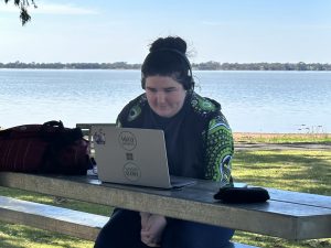 Tully sits at picnic table with laptop and headphones beside a lake