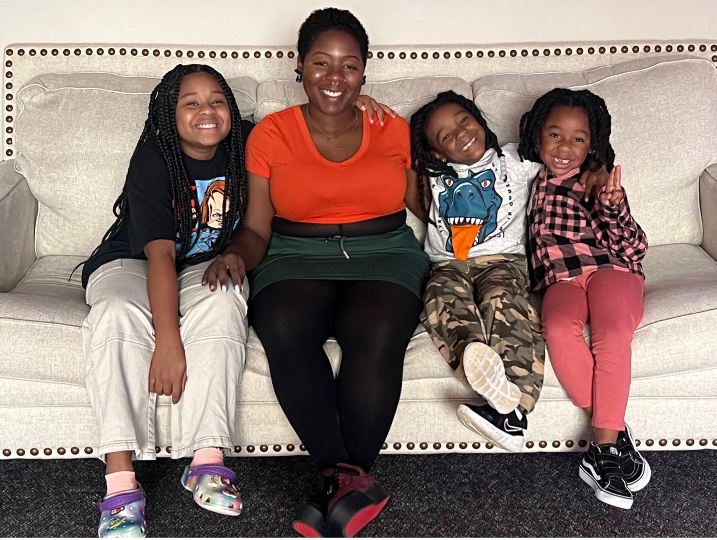 A dark-skinned woman with short, black hair and brown eyes wearing an orange shirt, dark green skirt, and black leggings sits on a cream-colored couch with one child on her left and two on her right, all with dark skin, dark hair and brown eyes, dressed casually in t-shirts and pants. 