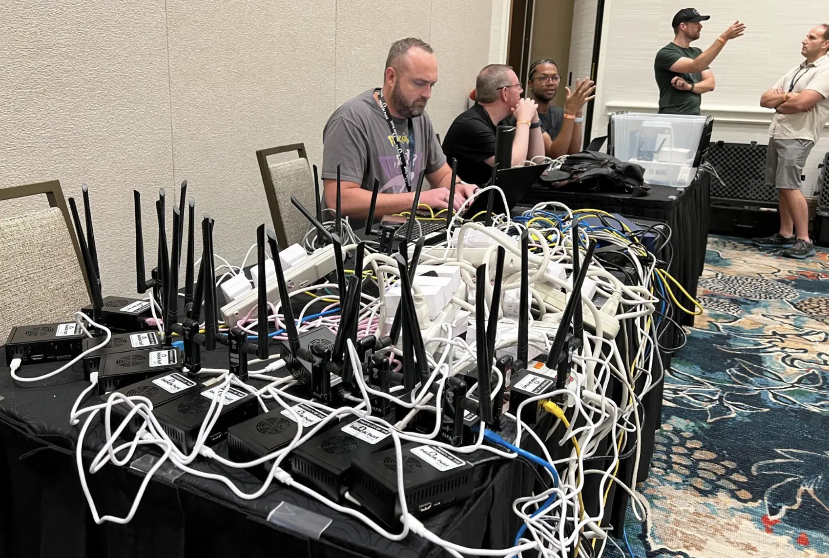 Man working at a table in a conference room; the table is completely covered in cords and power strips.