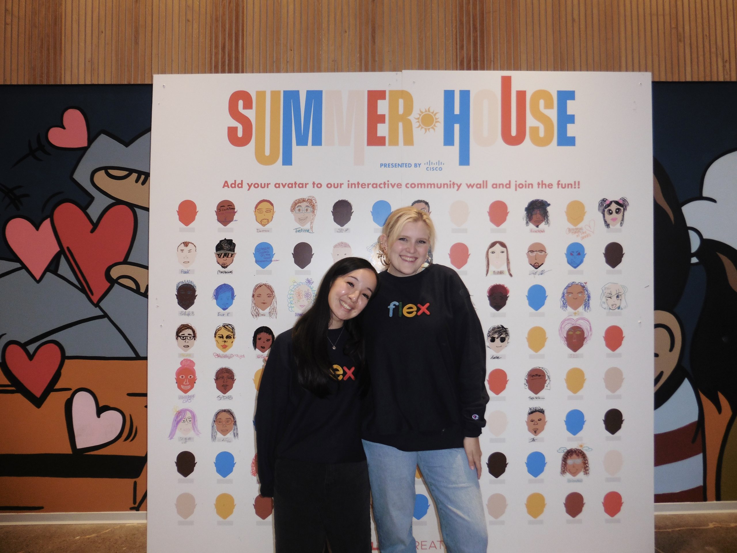Two young women, Micaela [blond] and her friend from the Flex program, Kathy [brunette], are smiling and standing in front of a graphic “SUMMER HOUSE presented by Cisco" sign. The sign features a grid of illustrated avatars with various faces. Both women are wearing dark sweatshirts with the word "flex" in colorful letters.
